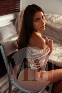 London escort Gloria sat on a chair wearing silky PJs