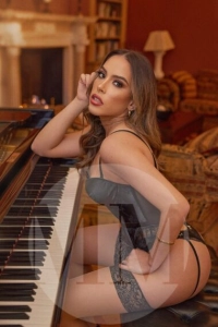 London escort Lilly sat next to a piano wearing black lingerie